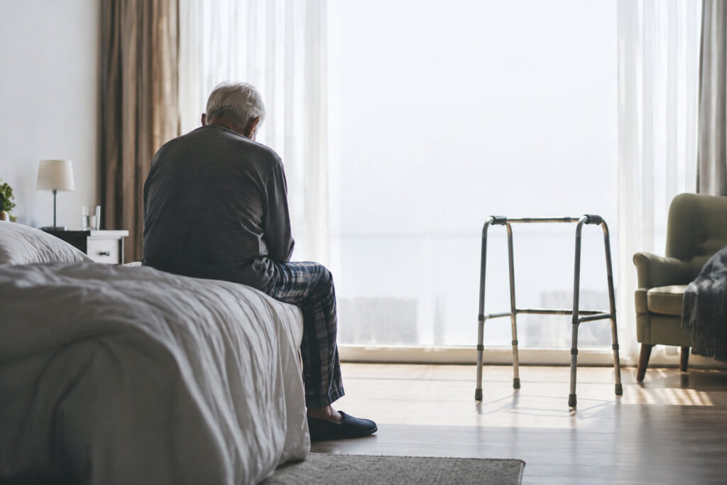 Un homme âgé aux cheveux gris assis sur le bord de son lit, de dos, avec un déambulateur à ses côtés dans une chambre.