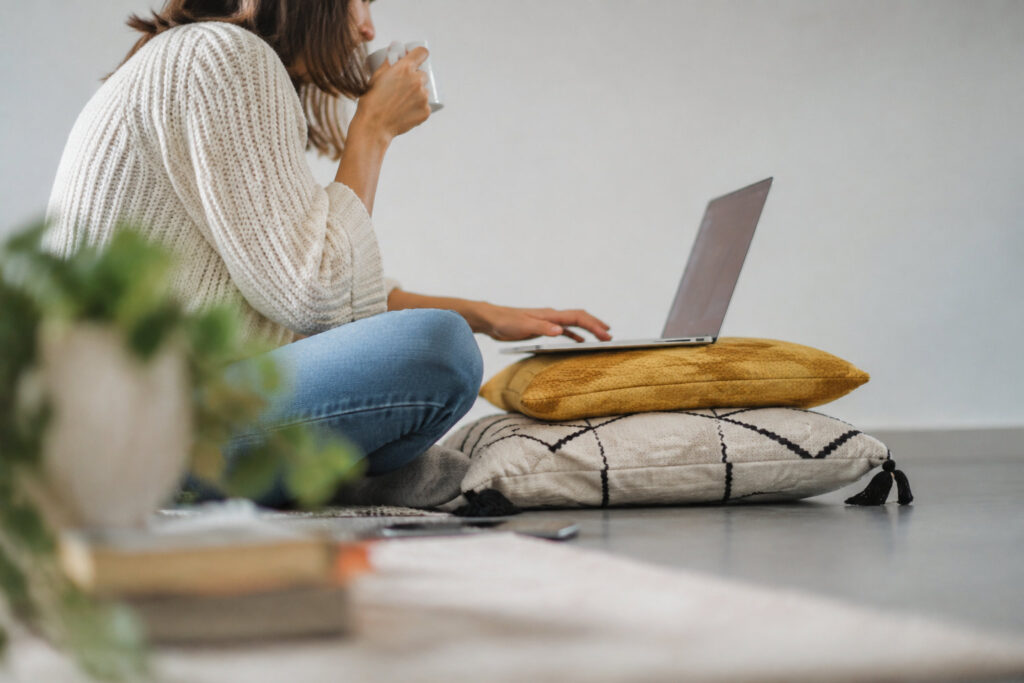 Une femme assise au sol utilisant son ordinateur portable avec une tasse à la main pour comparer sa mutuelle santé.